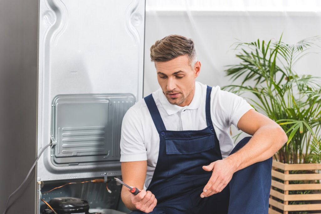 confused adult repairman looking at screwdriver while repairing refrigerator