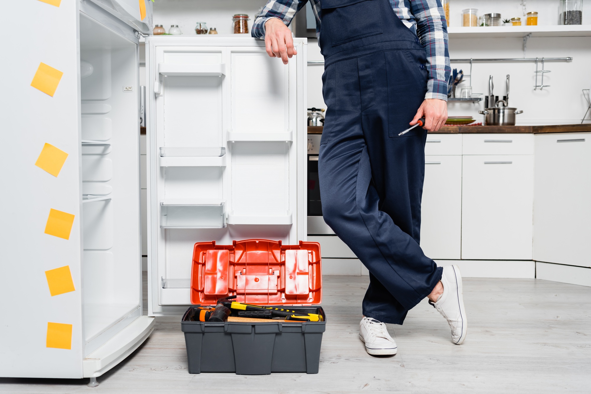 cropped view of young handyman with screwdriver leaning on fridge near toolbox in kitchen