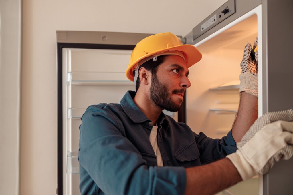 Male technician in uniform with screwdriver repairing refrigerator in kitchen