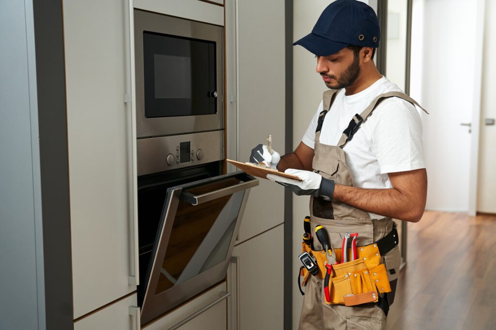 Professional service worker in uniform standing near oven and writing on clipboard in kitchen